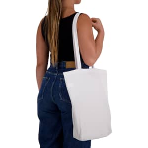 A white cotton long handle bag is slung over the shoulder of a person wearing a black top and denim jeans.