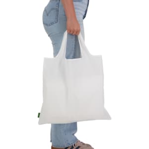 A white reusable grocery bag is being held by a person wearing blue jeans and shoes. The bag features a logo.