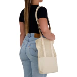 A tote bag in cream with a mesh design is held by a person wearing a black top and blue jeans.