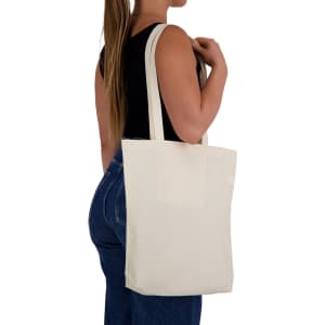 A cream tote bag held by a person, featuring shoulder straps and a simple design. The bag has a logo.