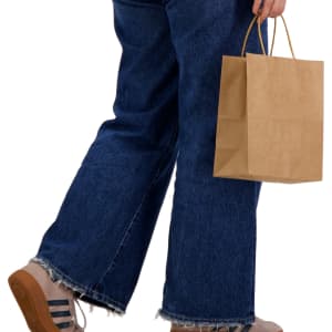 A medium brown paper bag held by a person wearing blue jeans and casual shoes. The bag has handles for easy carrying.