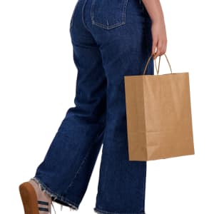 A person in blue jeans holds a large brown paper bag with handles, standing in a casual pose.