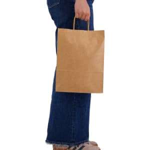 A large brown paper bag held by a person wearing blue jeans and casual shoes. The bag has handles and a simple design.
