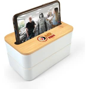 A white and bamboo lunch box with a phone holder lid, featuring a logo, displaying a phone on top.