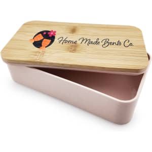 A pink lunch box with a bamboo lid featuring a logo, open to reveal a divided interior.