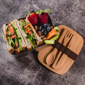 A bamboo lunch box featuring wraps, strawberries, blueberries, celery, and utensils, displayed on a textured surface.