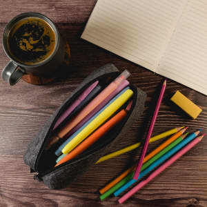 A gray felt utility case filled with colorful pens and pencils, alongside a notebook and a mug of coffee.