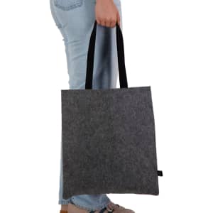 A grey felt tote bag with black handles being held by a person wearing light jeans. The bag has a logo.