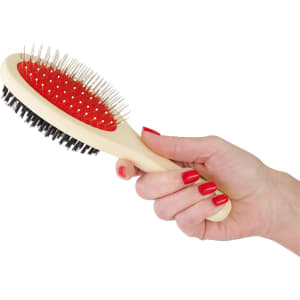 A pet grooming brush with a cream handle, red bristles, and black rubber base held in a hand with red nails.