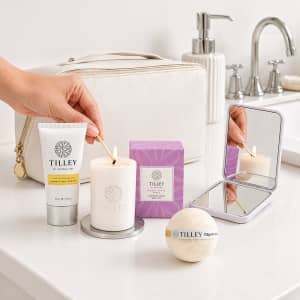 A cream toiletry bag with a candle, bath bomb, hand cream, and a compact mirror, arranged on a bathroom countertop.
