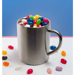 A stainless steel mug filled with assorted colour mini jelly beans, with some jelly beans scattered around the base.