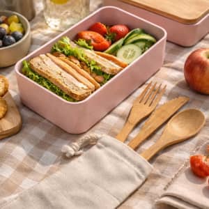 A bamboo cutlery set in a calico pouch, featuring forks and spoons, alongside a pink lunchbox with vegetables and fruit.