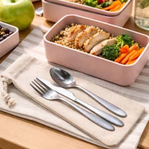 A cutlery set in silver with a calico pouch, placed beside a meal in a pink container on a striped tablecloth.