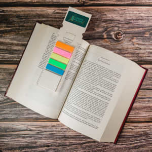 A rectangular bookmark in pastel colours rests in an opened book, featuring a logo at the top.