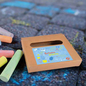 A box of Sketch Pavement Chalk with a colourful design, surrounded by green, pink, and yellow chalk pieces on pavement.