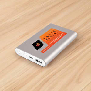 A silver power bank with a logo and an orange accent, featuring a USB port and indicator lights on a wooden surface.