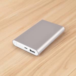 A silver power bank with a USB port on a wooden surface, featuring a sleek, minimal design and a logo.