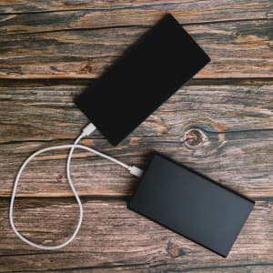 A black power bank rests on a wooden surface, connected by a charging cable to another black power bank.