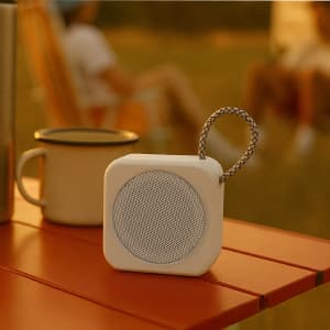 A compact white speaker with a woven strap, placed on a wooden table near a drink bottle.