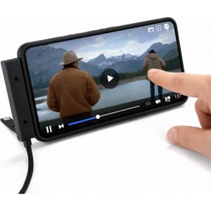 A smartphone displaying a video of a person by a lake, resting on a stand with a charging cable attached.