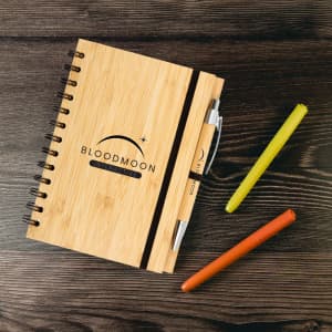 A bamboo notebook with a spiral binding, accompanied by a pen and three colored markers: yellow, orange, and green.