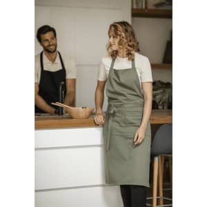A green recycled apron is displayed on a woman standing by a kitchen counter, with a man in an apron in the background.