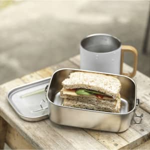 A stainless steel lunch box with a sandwich inside, placed beside a drink bottle on a wooden surface.