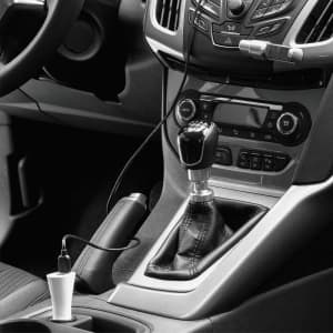 A dual car charger in white, plugged into a vehicle's centre console, next to the gear shift and dashboard controls.