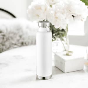 A drink bottle in white with a stainless steel base and cap, beside a flower arrangement on a table.