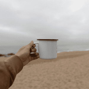 A drink bottle featuring a white enamel metal cup with a black rim, held against a beach backdrop.