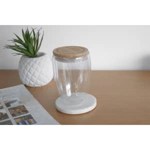 A drink bottle made of clear glass with a bamboo lid, set on a white marble base, alongside a decorative plant.