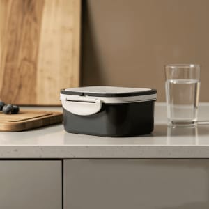 A black and white plastic lunch box on a kitchen countertop, beside a glass of water and a wooden cutting board.
