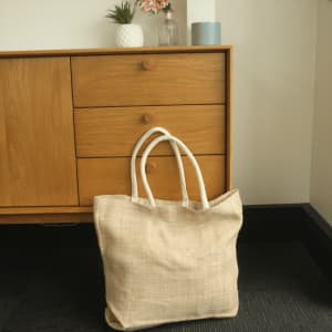 A herringbone patterned jute tote bag in natural and cream colours, with sturdy white handles, placed beside a wooden cabinet.
