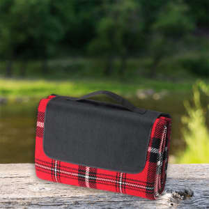A red and black checked picnic rug with a black handle, resting on a wooden surface near water.