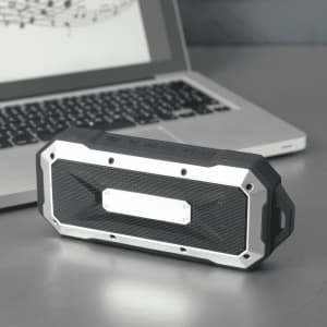A rugged black and silver waterproof Bluetooth speaker placed beside a laptop on a metallic surface.