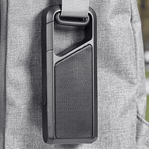 A portable black Bluetooth speaker attached to a grey bag with a strap. It has a textured surface and a prominent speaker grille.