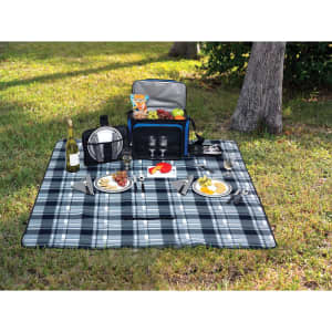 A picnic rug in shades of blue and grey, laid out with plates, glasses, cutlery, and a picnic basket on grass.