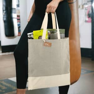 A beige and cream tote bag with a logo, held by a person, containing various items inside.