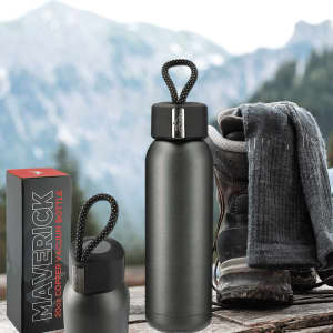 A drink bottle in matte black with a metallic lid, accompanied by a box and outdoor gear against a mountain backdrop.