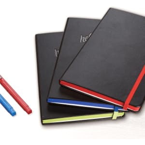 A set of three black bound journals with coloured elastic bands and pens in blue, red, and green.