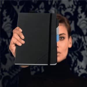 A black notebook held by a person, featuring a coloured tab and elastic strap, against a patterned background.