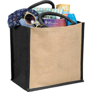 A jute tote bag featuring a natural canvas front and black sides, filled with sunglasses, a scarf, magazines, and other items.