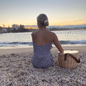 A wicker cooler basket in a light colour, positioned near a person on a beach at sunset. The scene features water and a cityscape.