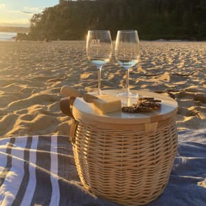 A wicker cooler basket in natural colour sits on a beach, with two wine glasses and a cheese board on top.