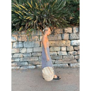 A wicker cooler basket in a light colour, carried by a person in a long, patterned dress against a stone wall backdrop.