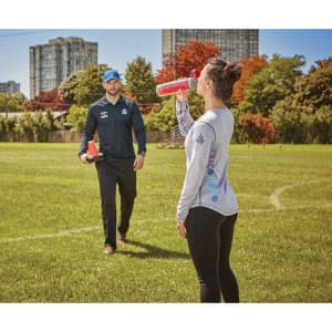 A woman in a light grey long sleeve polo shirt drinks from a red bottle while standing on a grassy field.