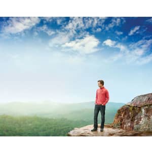 A man wearing a red quarter zip jumper stands on a rocky outcrop, overlooking a lush green landscape under a blue sky.