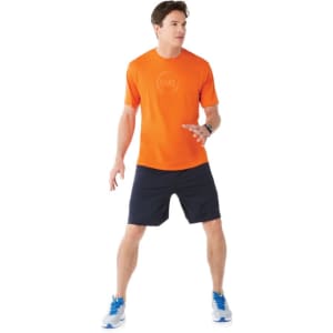 An orange short sleeve tech tee is worn by a man paired with dark shorts and blue athletic shoes. The shirt has a logo.