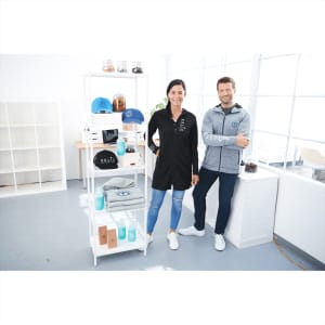A man in a grey knit zip hoody and a woman in a black shirt stand by a display of products, including caps and drink bottles.