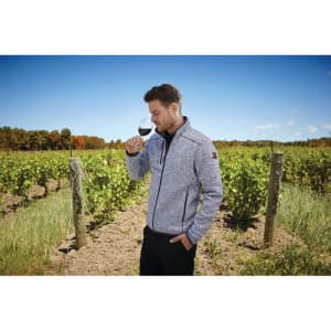 A man in a grey knit jacket poses with a wine glass in a vineyard setting, with green vines and blue sky in the background.